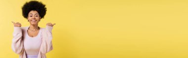african american woman in cardigan and top pointing with thumbs and smiling at camera isolated on yellow, banner