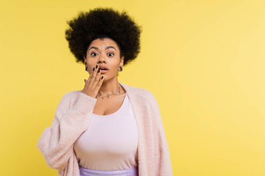 astonished african american woman in top and soft cardigan covering mouth with hand isolated on yellow