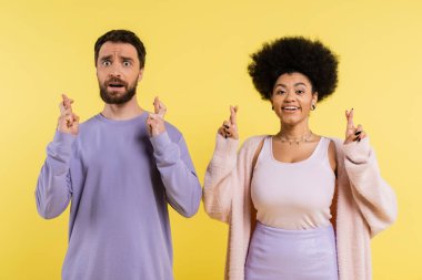 worried bearded man and optimistic african american woman crossing fingers isolated on yellow