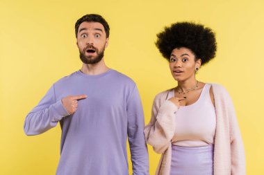 surprised interracial couple looking at camera and pointing with fingers at themselves isolated on yellow