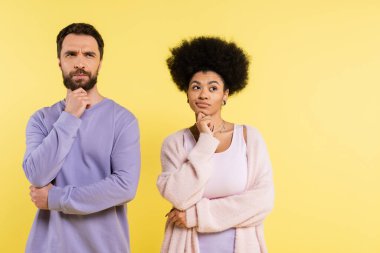 thoughtful man and african american woman holding hands near face and looking away isolated on yellow