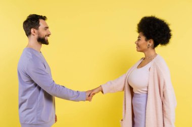 side view of bearded man and curly african american woman shaking hands and smiling at each other isolated on yellow