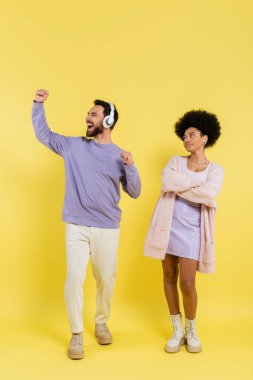 full length of excited man in headphones rejoicing near skeptical african american woman standing with crossed arms on yellow 