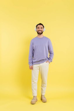 full length of trendy and happy man in wireless headphones standing with hand in pocket on yellow 