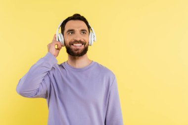 happy bearded man adjusting wireless headphones while listening music isolated on yellow