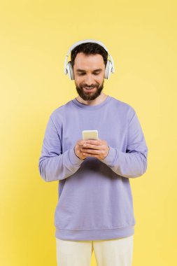smiling bearded man in purple pullover and wireless headphones using smartphone isolated on yellow