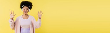 joyful african american woman listening music in wireless headphones and waving hands isolated on yellow, banner