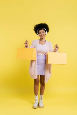 full length of happy african american woman in wireless headphones and boots holding shopping bags on yellow background