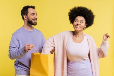 happy and curious african ametrican woman checking shopping bag near smiling bearded man isolated on yellow