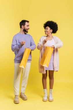 full length of cheerful interracial couple looking at each other while standing with shopping bags on yellow 