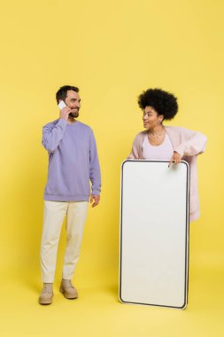 african american woman pointing at phone template near smiling bearded man talking on mobile phone on yellow background