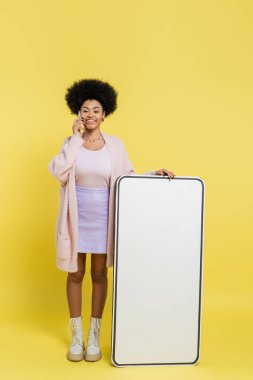 full length of joyful african american woman talking on cellphone near blank mock-up of mobile phone on yellow background