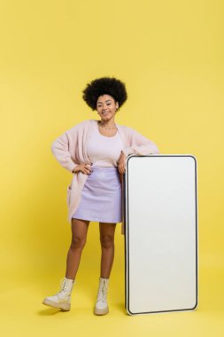 full length of happy african american woman standing with hand on hip near carton template of smartphone on yellow background