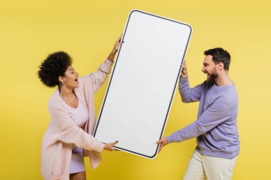 side view of thrilled interracial couple screaming while holding huge phone template isolated on yellow clipart