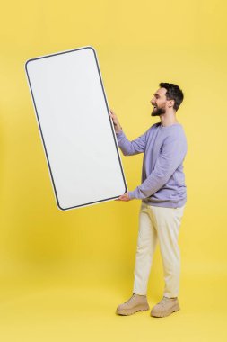 side view of happy bearded man holding huge template of mobile phone on grey background