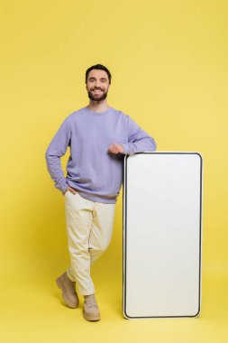 full length of happy bearded man posing with hand in pocket near white mock-up of mobile phone on grey background