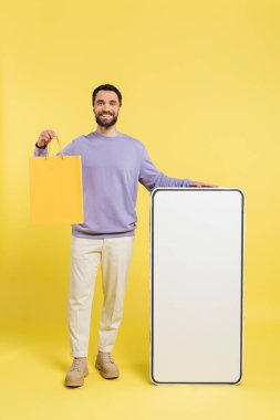 full length of cheerful bearded man standing with shopping bag and huge template of smartphone on grey background