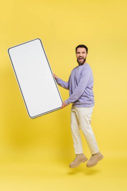 full length of excited bearded man levitating with huge mobile phone template on grey background