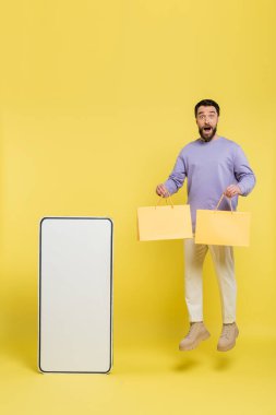 full length of excited man levitating with shopping bags near carton template of mobile phone on grey background