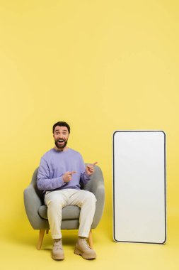 full length of astonished man sitting in armchair and pointing at big phone mockup on grey background