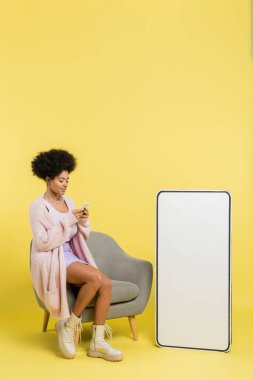 full length of stylish african american woman using mobile phone on armchair near white template of cellphone on yellow background