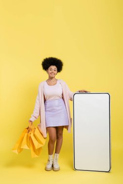 full length of joyful and trendy african american woman standing with shopping bags near carton model of smartphone on yellow background