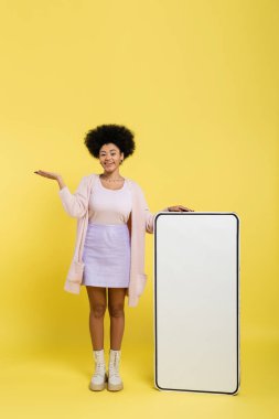 full length of cheerful african american woman pointing with hand near huge template of mobile phone on yellow background