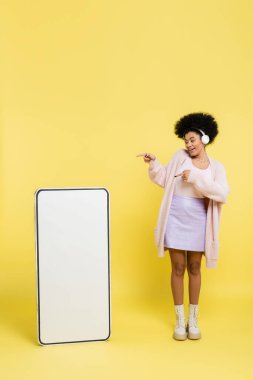 full length of happy african american woman in wireless headphones pointing at empty template of cellphone on yellow background