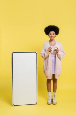 full length of pleased african american woman standing with wireless headphones near white mock-up of cellphone on yellow background