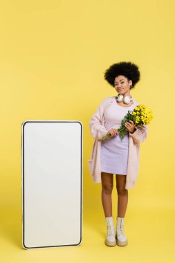 full length of african american woman with wireless headphones and flowers smiling near blank template of smartphone on yellow 
