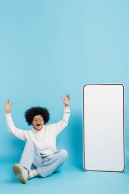 full length of excited african american woman in hoodie and eyeglasses waving hands while sitting near phone template on blue 