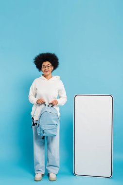full length of african american student holding backpack and looking at camera near carton phone mock-up on blue background