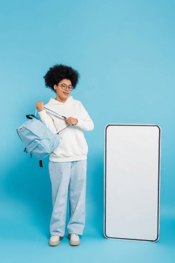 full length of smiling african american student with backpack looking at camera near empty phone template on blue background