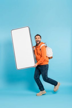 full length of excited student with backpack carrying huge mockup of smartphone on blue background