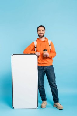 full length of bearded student with takeaway drink smiling at camera near white phone mock-up on blue background