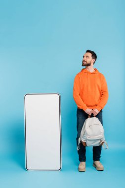 full length of bearded student in orange hoodie standing with backpack near huge phone template on blue background