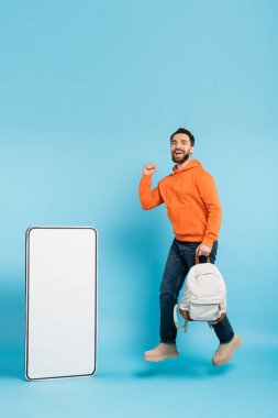 full length of cheerful bearded student with backpack showing win gesture while levitating near phone template on blue background