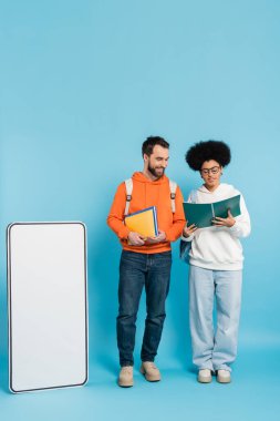 full length of african american woman looking in copybook near smiling student and carton smartphone template on blue background