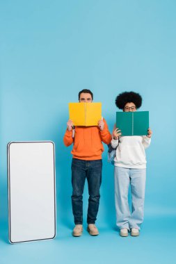 full length of interracial students obscuring face with notebooks near blank template of mobile phone on blue background