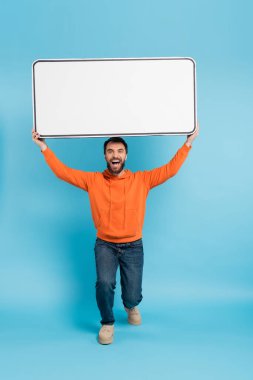 full length of excited man in orange hoodie and jeans holding big phone template above head on blue background