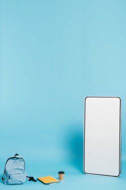 vertical view of white phone template near backpack and notebooks with paper cup on blue background