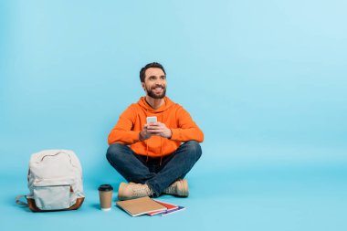 bearded student with mobile phone sitting near notebooks and backpack while looking away on blue background clipart