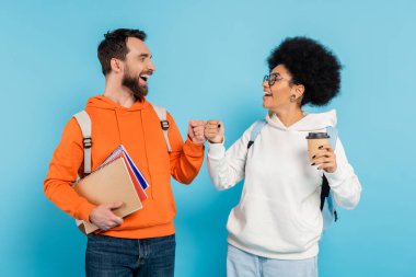 cheerful interracial students with coffee to go and notebooks doing fist bump isolated on blue