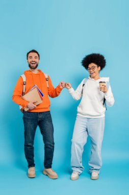 full length of multiethnic students with notebooks and takeaway drink doing fist bump while smiling at camera on blue background
