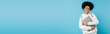 positive african american student with curly hair and eyeglasses holding laptop isolated on blue, banner