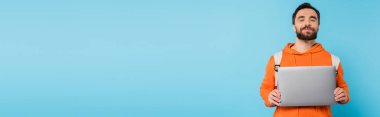 smiling bearded student with closed eyes holding laptop isolated on blue, banner