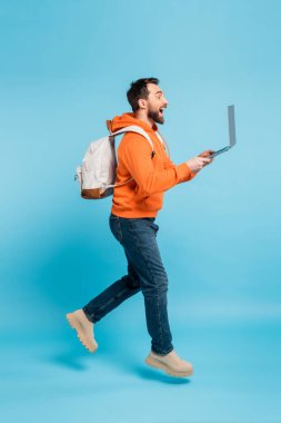 side view of excited student with backpack and laptop levitating on blue background