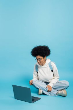 full length of african american student in eyeglasses and white hoodie sitting with crossed legs near laptop on blue background