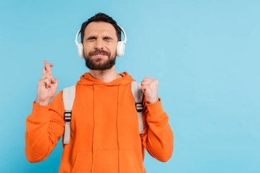positive bearded student in wireless headphones crossing fingers while standing with closed eyes isolated on blue