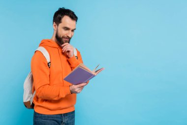 thoughtful and positive student with backpack reading book and touching beard isolated on blue